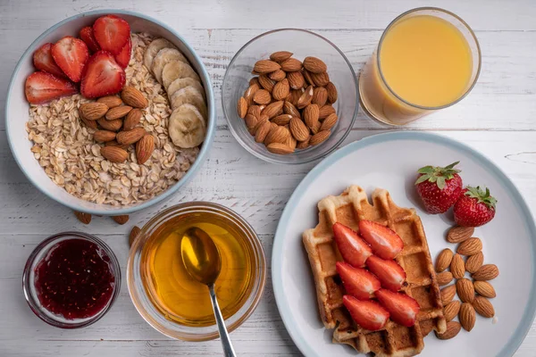 Breakfast menu food top view. Oatmeal, waffles, berry, nuts, cereal, honey, orange juice, strawberry jam on a white table. Delicious menu. Dessert. Ingredients