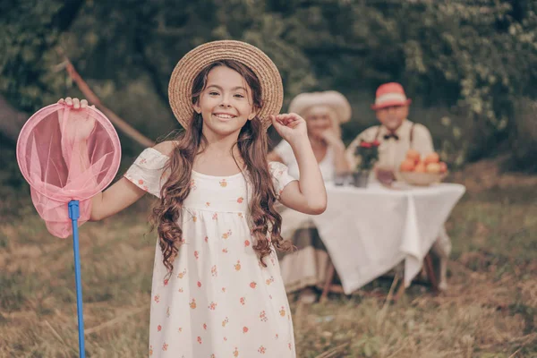 穿着太阳衣 头戴一顶蝴蝶网的帽子 快乐的小女孩笑着 一个孙女在花园里的画像 在背景中 祖父母坐在桌旁 在公园里喝柠檬水和水果 — 图库照片