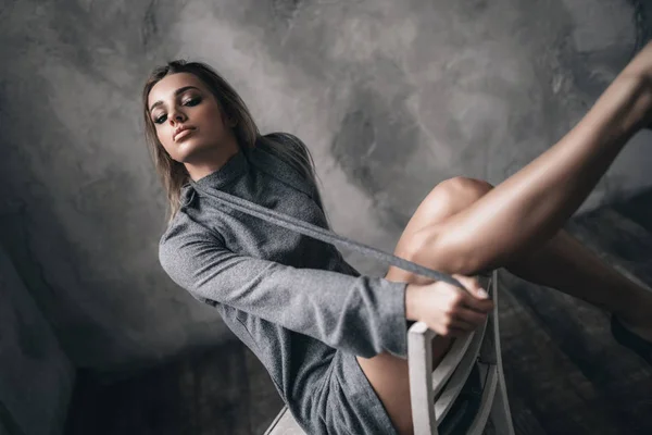 椅子の屋内スタジオで夜のドレス姿でポーズをとる長い脚を持つ魅力的なセクシーな女の子 官能的な魅力的なブロンド女性モデル見ます上のカメラ上の灰色の壁の背景 — ストック写真