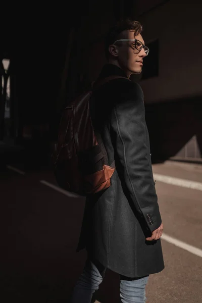 Fashion man portrait from behind. A young guy in a black coat, glasses,jeans and a backpack stands against the background of the tunnel