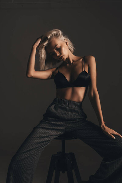 Fashion young beauty woman with blonde hair in lingerie and classic trousers posing on chair on black background in room