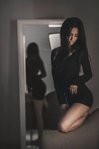 Seductive woman portrait of trendy woman in black dress on armchair. Stylish fashion girl model posing near mirror in bedroom
