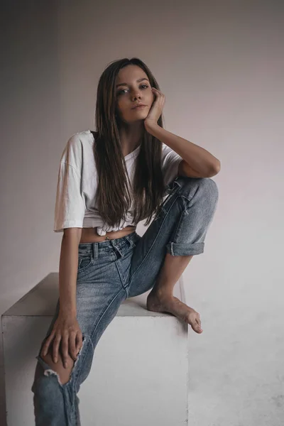 Sexy hot woman in shirt, jeans and sneakers posing in studio on cube on white background. Seductive glamour young girl model portrait.