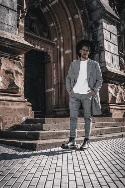 Stijlvol jongeman portret in stedelijke achtergrond. Knappe man met baard model wandelen buiten in casual jas met stijlvolle hoed en kapsel in de stad straat — Stockfoto