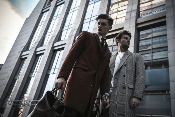Two fashion men models posing on city street. Wearing in classic and casual closes. Suit, coat, shirt, sweater, boots and leather bag. Men model test near business center