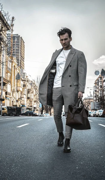 Fashion model man walking on road near business center. Street photo shooting. Gay wearing in gray coat, white sweater, black shoes, brown leather bag.