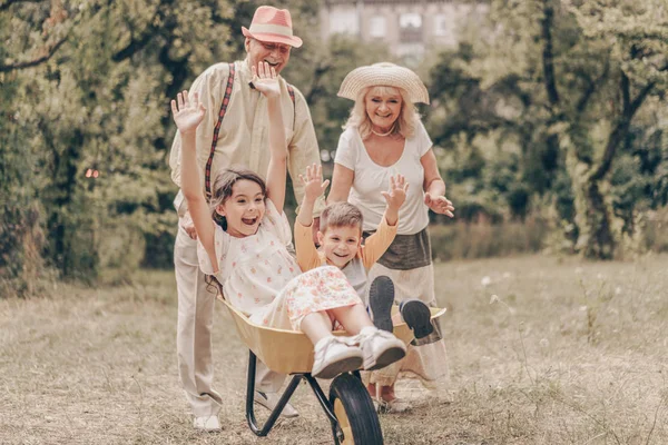 祖父母在公园和孙子孙女一起玩推车 一家人在花园里玩得很开心 老年人很高兴见到孙女和孙子 图库图片