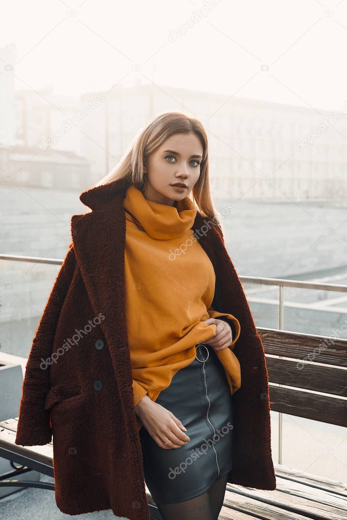 A beautiful young girl blonde walks through the streets of the city, smiling, waving her coat. She is wearing an orange sweater and black tights, a red coat. Street casual style. Emotion of joy