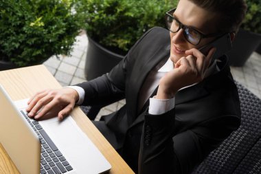 Her zaman müsait. Güzel genç iş adamı dizüstü bilgisayarıyla çalışıyor ve dışarıda masada otururken telefonla konuşuyor.