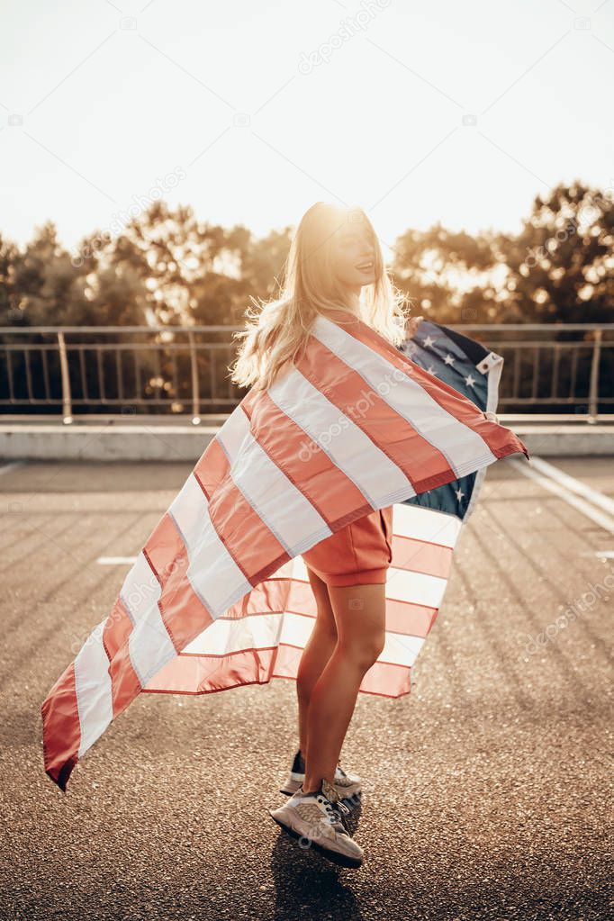 Beauty portrait of active happy girl with american flag on road near parking in summer sunset in evening. Stylish urban woman model posing in hoodie