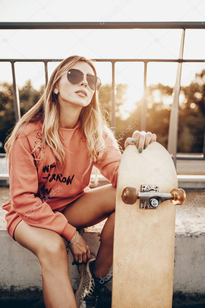 Beauty portrait of active girl with skateboard on road near parking in summer sunset in evening. Stylish urban woman model posing in red hoodie, sneakers