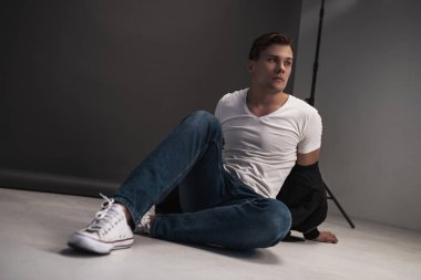 Stüdyoda gri arka planda poz veren şık bir genç. Beyaz gömlek, kot pantolon ve beyaz spor ayakkabı giymişti.