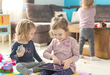 İki küçük kız kardeş evde tablet kullanmak ve oyuncaklar ortasında otur. Komik bir şey izliyor.