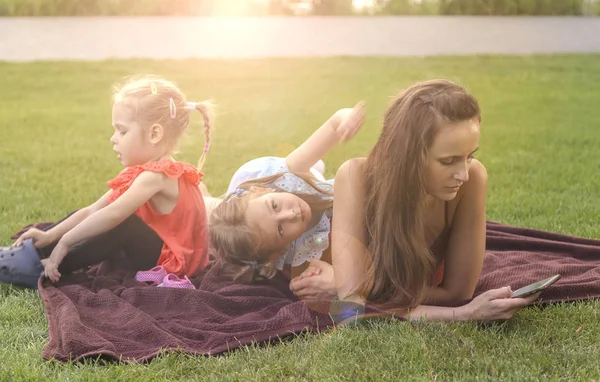 Madre con hijas de picnic por el río, ella chats en el teléfono inteligente — Foto de Stock