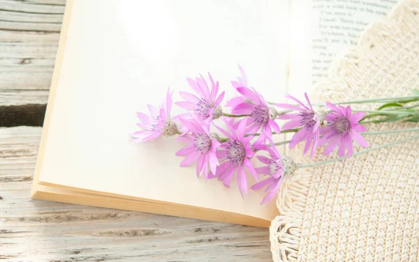 open book with beautiful delicate flowers in the fields of a hat lie on a wooden table in the garden. English text on the pages of the book is changed, letters are partially erased.