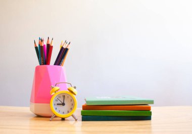 Beyaz bir duvara karşı renkli kalemler, defterler ve çalar saat.