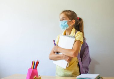Yüzünde koruyucu bir tıbbi maske takan okul kızı elinde okul malzemelerini tutuyor.