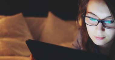 Geceleri Tablet Bilgisayar kullanan, gözlüklü bir kadın. Yavaş Hareket 4K DCI, Yaklaş. Genç iş kadını geç saatlere kadar çalışıyor, yatakta yatıyor. Günlük yaşamda teknoloji.