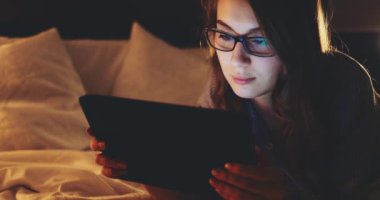 Geceleri Tablet Bilgisayar kullanan, gözlüklü bir kadın. Yavaş Hareket 4K DCI, Yaklaş. Genç iş kadını geç saatlere kadar çalışıyor, yatakta yatıyor. Günlük yaşamda teknoloji.