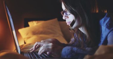 Gece Evde Dizüstü bilgisayarda çalışan bir kadın. 4K Yavaşlama Hareketi, DCI. Genç bir kadın, gözlük takıyor, yatağa uzanıyor ve taşınabilir bilgisayar kullanıyor. Günlük yaşamda teknoloji.