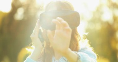Sanal Gerçeklik Gözlüklü Kadın Tezahürat Yapıyor. Yavaş haraket 4K. Sanal 3 boyutlu dünyanın tadını çıkaran, VR kulaklıkla oynayan neşeli genç bir kadın. Güneşli alan arkaplanı, hayali gerçekliğin içinde.