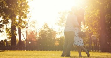 Şehir parkında el ele tutuşup sohbet eden genç mutlu çift. 4K DCI Yavaş Hareketi. Güzel, romantik, gülümseyen bir kadın ve bir erkek gün batımında kameraya doğru yürüyorlar.