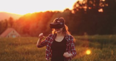 Sanal Gerçeklik Gözlüklü Kadın Tezahürat Yapıyor. Yavaş haraket 4K. Sanal 3 boyutlu dünyanın tadını çıkaran, VR kulaklıkla oynayan neşeli genç bir kadın. Güneşli alan arkaplanı, hayali gerçekliğin içinde.