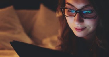 Geceleri Tablet Bilgisayar kullanan, gözlüklü bir kadın. Yavaş Hareket 4K DCI, Yaklaş. Genç iş kadını geç saatlere kadar çalışıyor, yatakta yatıyor. Günlük yaşamda teknoloji.