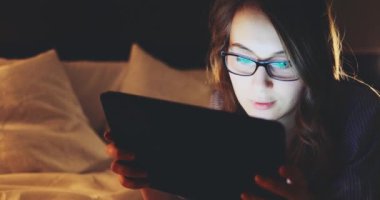 Geceleri Tablet Bilgisayar kullanan, gözlüklü bir kadın. Yavaş Hareket 4K DCI, Yaklaş. Genç iş kadını geç saatlere kadar çalışıyor, yatakta yatıyor. Günlük yaşamda teknoloji.