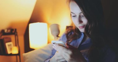 Akşamları Evde Akıllı Telefon Kullanan Kadın. Yavaş hareket et. 4K DCI. Sosyal medya uygulaması kullanan, sohbet eden, yatakta uzanan genç bir kadın. Günlük yaşamda teknoloji.