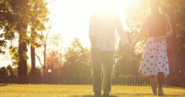 Şehir parkında el ele tutuşup sohbet eden genç mutlu çift. 4K DCI Yavaş Hareketi. Güzel, romantik, gülümseyen bir kadın ve bir erkek gün batımında kameraya doğru yürüyorlar.