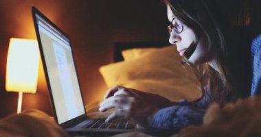Gece Evde Dizüstü bilgisayarda çalışan bir kadın. 4K Yavaşlama Hareketi, DCI. Genç bir kadın, gözlük takıyor, yatağa uzanıyor ve taşınabilir bilgisayar kullanıyor. Günlük yaşamda teknoloji.