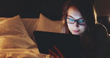 Geceleri Tablet Bilgisayar kullanan, gözlüklü bir kadın. Yavaş Hareket 4K DCI, Yaklaş. Genç iş kadını geç saatlere kadar çalışıyor, yatakta yatıyor. Günlük yaşamda teknoloji.