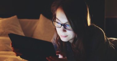 Geceleri Tablet Bilgisayar kullanan, gözlüklü bir kadın. Yavaş Hareket 4K DCI, Yaklaş. Genç iş kadını geç saatlere kadar çalışıyor, yatakta yatıyor. Günlük yaşamda teknoloji.