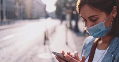 Akıllı Telefon kullanan koruyucu maskeli genç bir kadın güneşli bir sabah şehrinde tramvayı bekliyor. Yavaş hareket, Gimbal Dengeleyici. Hava kirliliği ve koronavirüse karşı maskeli iş kadını nakil aracı kullanıyor.