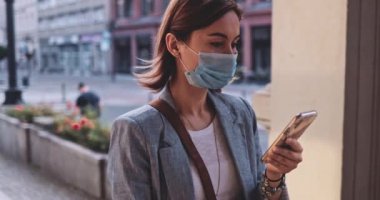 Şehirde koruyucu maske takan, akıllı telefon kullanan bir kadın var. Yavaş hareket, Gimbal Dengeleyici. Hava kirliliği ve Coronavirus Covid-19 'a karşı maskeli genç bir kadın. Şehir Sabahı.