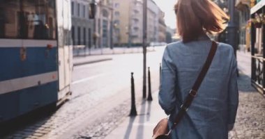 Koruyucu Maskeli Genç Kadın Güneşli Sabah Şehrinde Tramvay Bekliyor. Yavaş hareket, Gimbal Dengeleyici. Hava kirliliği ve koronavirüse karşı maskeli iş kadını toplu taşıma kullanıyor.