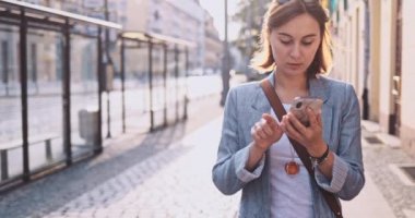 Genç İşkadını Akıllı Telefon Kullanıyor, Güneşli Sabah Şehrinde İşe Gidiyor. Yavaş hareket, Gimbal Dengeleyici. Şehirde cep telefonu olan çekici, profesyonel bir iş kadını. Mercek Fişeği.
