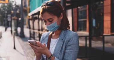 Akıllı Telefon kullanan koruyucu maskeli genç bir kadın güneşli bir sabah şehrinde tramvayı bekliyor. Yavaş hareket, Gimbal Dengeleyici. Hava kirliliği ve koronavirüse karşı maskeli iş kadını nakil aracı kullanıyor.
