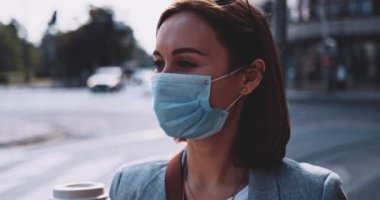 Koruyucu maskeli bir kadın kahve içiyor, şehirde yürüyor. Yavaş bir hareket yap. Genç bayan hava kirliliğine ve Coronavirus covid-19 'a karşı maskesini çıkararak kağıt bardaktan su içiyor..