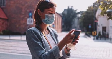 Güneşli Sabah Şehrinde Koruyucu Maskeli Genç Kadın Dezenfekte Etme Akıllı Telefonu. Yavaş hareket, Gimbal Dengeleyici. Hava kirliliğine karşı maskeli iş kadını ve koronavirüs telefon ekranını sterilize ediyor