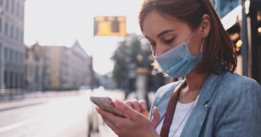Akıllı Telefon kullanan koruyucu maskeli genç bir kadın güneşli bir sabah şehrinde tramvayı bekliyor. Yavaş hareket, Gimbal Dengeleyici. Hava kirliliği ve koronavirüse karşı maskeli iş kadını nakil aracı kullanıyor.