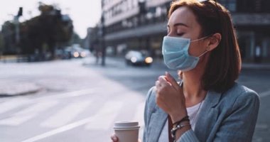 Koruyucu maskeli bir kadın kahve içiyor, şehirde yürüyor. Yavaş bir hareket yap. Genç bayan hava kirliliğine ve Coronavirus covid-19 'a karşı maskesini çıkararak kağıt bardaktan su içiyor..