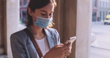 Şehirde koruyucu maske takan, akıllı telefon kullanan bir kadın var. Yavaş hareket, Gimbal Dengeleyici. Hava kirliliği ve Coronavirus Covid-19 'a karşı maskeli genç bir kadın. Şehir Sabahı.