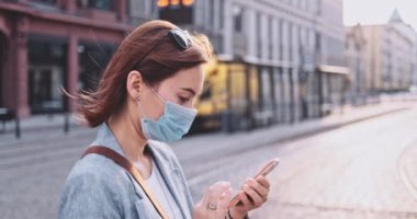 Şehirde koruyucu maske takan, akıllı telefon kullanan bir kadın var. Yavaş hareket, Gimbal Dengeleyici. Hava kirliliği ve Coronavirus Covid-19 'a karşı maskeli genç bir kadın. Şehir Sabahı.