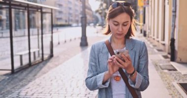 Genç İşkadını Akıllı Telefon Kullanıyor, Güneşli Sabah Şehrinde İşe Gidiyor. Yavaş hareket, Gimbal Dengeleyici. Şehirde cep telefonu olan çekici, profesyonel bir iş kadını. Mercek Fişeği.