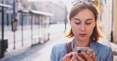 Genç İşkadını Akıllı Telefon Kullanıyor, Güneşli Sabah Şehrinde İşe Gidiyor. Yavaş hareket, Gimbal Dengeleyici. Şehirde cep telefonu olan çekici, profesyonel bir iş kadını. Mercek Fişeği.