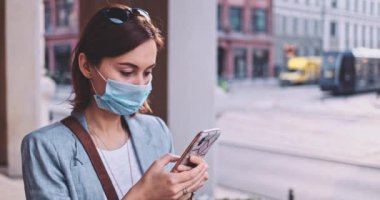 Şehirde koruyucu maske takan, akıllı telefon kullanan bir kadın var. Yavaş hareket, Gimbal Dengeleyici. Hava kirliliği ve Coronavirus Covid-19 'a karşı maskeli genç bir kadın. Şehir Sabahı.