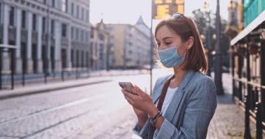 Akıllı Telefon kullanan koruyucu maskeli genç bir kadın güneşli bir sabah şehrinde tramvayı bekliyor. Yavaş hareket, Gimbal Dengeleyici. Hava kirliliği ve koronavirüse karşı maskeli iş kadını nakil aracı kullanıyor.