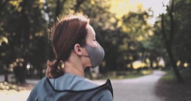 Güneşli şehir parkında koşan koruyucu maske takan bir kadın dışarıda egzersiz yapıyor. Gimbal sabitlendi, yavaşlama hareketi yapıldı. Hava kirliliğine karşı maskeli sporcu kadın ve Covid19 Sabah Koşusu.